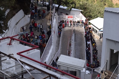 A large crowd of people is gathered in a narrow outdoor area that appears to be a queue. They are dressed warmly, suggesting cold weather, with snow visible on the ground and nearby surfaces. The scene is bordered by white structures and some greenery in the background.