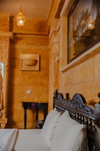 Ornately detailed interior featuring carved wooden furniture, patterned walls, and a hanging lantern casting a warm glow. A bed with plush pillows is positioned in the foreground, and an artistic framed picture is mounted on the wall.