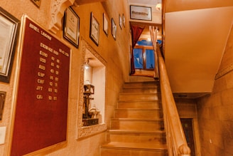 A warmly lit indoor staircase with polished wooden steps leads to a window adorned with red curtains. The walls display several framed certificates and a red board listing currency exchange rates. An ornate niche houses a small decorative item in a frame, further enhancing the space's cozy and elegant feel.