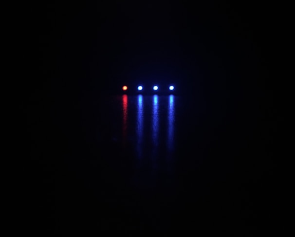 A dark background with five small, illuminated lights arranged horizontally; one red light followed by four blue lights casting reflections below them.