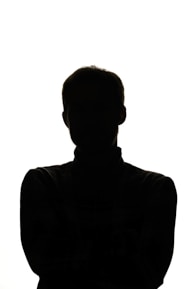 A silhouette of a person is set against a bright white background. The figure features a discernible head and shoulders, with arms crossed or tucked in front, making it instantly recognizable by its shape. The lighting emphasizes a stark contrast between the subject and the backdrop.