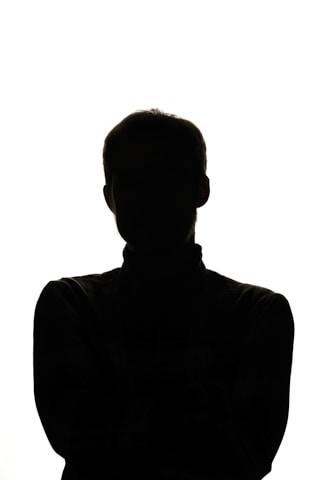 A silhouette of a person is set against a bright white background. The figure features a discernible head and shoulders, with arms crossed or tucked in front, making it instantly recognizable by its shape. The lighting emphasizes a stark contrast between the subject and the backdrop.