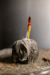 A carved elephant-shaped incense holder with a burning incense stick inserted. The smoke elegantly rises and swirls around the wooden figure, which is adorned with intricate carvings. The background is softly blurred, creating a serene and contemplative ambiance.