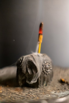 A carved elephant-shaped incense holder with a burning incense stick inserted. The smoke elegantly rises and swirls around the wooden figure, which is adorned with intricate carvings. The background is softly blurred, creating a serene and contemplative ambiance.