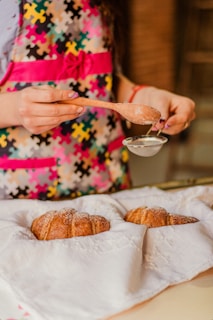 A person wearing a colorful apron with a puzzle pattern is sifting powdered sugar over two croissants resting on a white cloth. The scene suggests a homey and cozy kitchen environment.