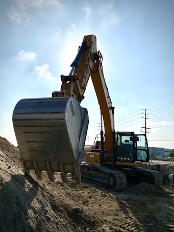 A large yellow excavator with a metallic bucket is positioned on a mound of dirt. The machine is situated outdoors under a blue sky with scattered clouds. There are electric poles and power lines in the background, as well as some industrial buildings.