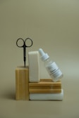 Several bars of soap are stacked with a pair of scissors inserted into one of them. A glass dropper bottle labeled 'The Ordinary' leans against the soap stack. The background is a plain beige color.