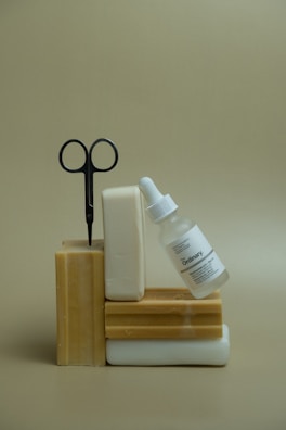 Several bars of soap are stacked with a pair of scissors inserted into one of them. A glass dropper bottle labeled 'The Ordinary' leans against the soap stack. The background is a plain beige color.