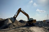 A large yellow excavator is stationed on a sandy construction site, with its arm extended and bucket poised over a mound of dirt. The scene is set under an expansive blue sky with a few scattered clouds, and there are traces of machinery tracks in the sand. Utility poles and industrial elements are visible in the background.