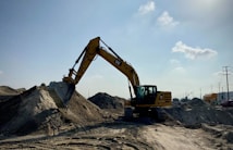 A large yellow excavator is stationed on a sandy construction site, with its arm extended and bucket poised over a mound of dirt. The scene is set under an expansive blue sky with a few scattered clouds, and there are traces of machinery tracks in the sand. Utility poles and industrial elements are visible in the background.