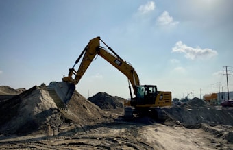 A large yellow excavator is stationed on a sandy construction site, with its arm extended and bucket poised over a mound of dirt. The scene is set under an expansive blue sky with a few scattered clouds, and there are traces of machinery tracks in the sand. Utility poles and industrial elements are visible in the background.