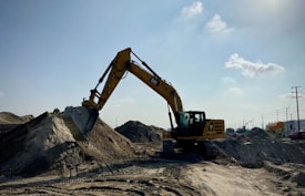 A large yellow excavator is stationed on a sandy construction site, with its arm extended and bucket poised over a mound of dirt. The scene is set under an expansive blue sky with a few scattered clouds, and there are traces of machinery tracks in the sand. Utility poles and industrial elements are visible in the background.
