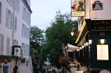 A quaint Parisian street features a charming café with outdoor seating on the right side, adorned with decorative lamps and a sign reading 'La Bonne Franquette'. The left side of the street is lined with a tall white building with numerous windows. Lush green trees frame the street further into the background. People are seen casually strolling and interacting near the café, adding to the scene's lively atmosphere.