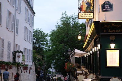 A quaint Parisian street features a charming caf&eacute; with outdoor seating on the right side, adorned with decorative lamps and a sign reading 'La Bonne Franquette'. The left side of the street is lined with a tall white building with numerous windows. Lush green trees frame the street further into the background. People are seen casually strolling and interacting near the caf&eacute;, adding to the scene's lively atmosphere.