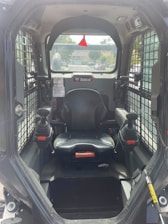 The interior of a Bobcat construction vehicle with a black seat and two joysticks on either side. The cabin is enclosed with a wire grill on the sides and a clear view towards the outside, showing a residential area in the background.