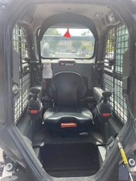 The interior of a Bobcat construction vehicle with a black seat and two joysticks on either side. The cabin is enclosed with a wire grill on the sides and a clear view towards the outside, showing a residential area in the background.