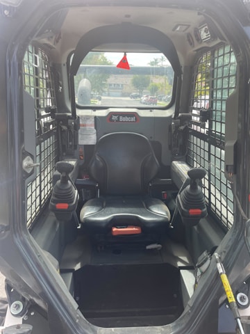 The interior of a Bobcat construction vehicle with a black seat and two joysticks on either side. The cabin is enclosed with a wire grill on the sides and a clear view towards the outside, showing a residential area in the background.