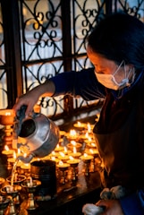 A person is pouring liquid from a kettle into small metal lamps, which are lit and arranged on a table. The lighting casts a warm glow, highlighting the activity. The person is wearing a mask and an apron, focusing on their task.