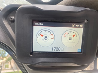 A digital screen displaying vehicle dashboard indicators. It shows two circular gauges; the left one depicts an engine temperature gauge with a water symbol, and the right one indicates a fuel gauge. The top left corner displays the hours used, reading 298.5 hrs, and the bottom shows a digital number 1720 with a lighting symbol beside it. The screen is surrounded by a black frame held in place by a bolt.