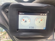 A digital screen displaying vehicle dashboard indicators. It shows two circular gauges; the left one depicts an engine temperature gauge with a water symbol, and the right one indicates a fuel gauge. The top left corner displays the hours used, reading 298.5 hrs, and the bottom shows a digital number 1720 with a lighting symbol beside it. The screen is surrounded by a black frame held in place by a bolt.