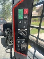 A control panel of machinery or a vehicle featuring various buttons and knobs with different functions. The panel includes symbols such as a stop button, headlight control, parking indicator, and others. It is bordered by a prominent red line, and the view outside shows greenery and a residential area.