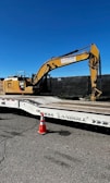 A large yellow excavator with 'Subzero Excavating' branding is parked on a flatbed trailer. There is a bright orange traffic cone positioned on the ground in front of the trailer, which is situated on a paved surface. A clear, deep blue sky is visible above, and a black fence runs behind the machinery.