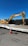 A large yellow excavator with 'Subzero Excavating' branding is parked on a flatbed trailer. There is a bright orange traffic cone positioned on the ground in front of the trailer, which is situated on a paved surface. A clear, deep blue sky is visible above, and a black fence runs behind the machinery.