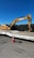 A large yellow excavator with 'Subzero Excavating' branding is parked on a flatbed trailer. There is a bright orange traffic cone positioned on the ground in front of the trailer, which is situated on a paved surface. A clear, deep blue sky is visible above, and a black fence runs behind the machinery.