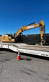 A large yellow excavator with 'Subzero Excavating' branding is parked on a flatbed trailer. There is a bright orange traffic cone positioned on the ground in front of the trailer, which is situated on a paved surface. A clear, deep blue sky is visible above, and a black fence runs behind the machinery.