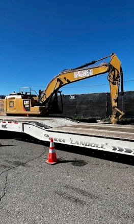 A large yellow excavator with 'Subzero Excavating' branding is parked on a flatbed trailer. There is a bright orange traffic cone positioned on the ground in front of the trailer, which is situated on a paved surface. A clear, deep blue sky is visible above, and a black fence runs behind the machinery.