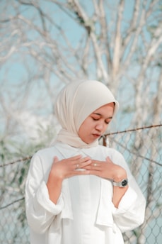 A person wearing a light-colored headscarf and a white flowing garment, standing with hands placed gently on their chest. The background features a blurred chain-link fence and bare trees, with a blue sky visible. The person appears contemplative or serene.