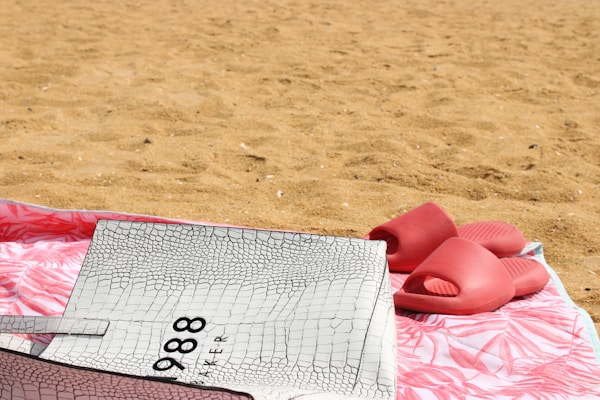 Casual white rubber sandals placed beside a beach towel on sandy shore.