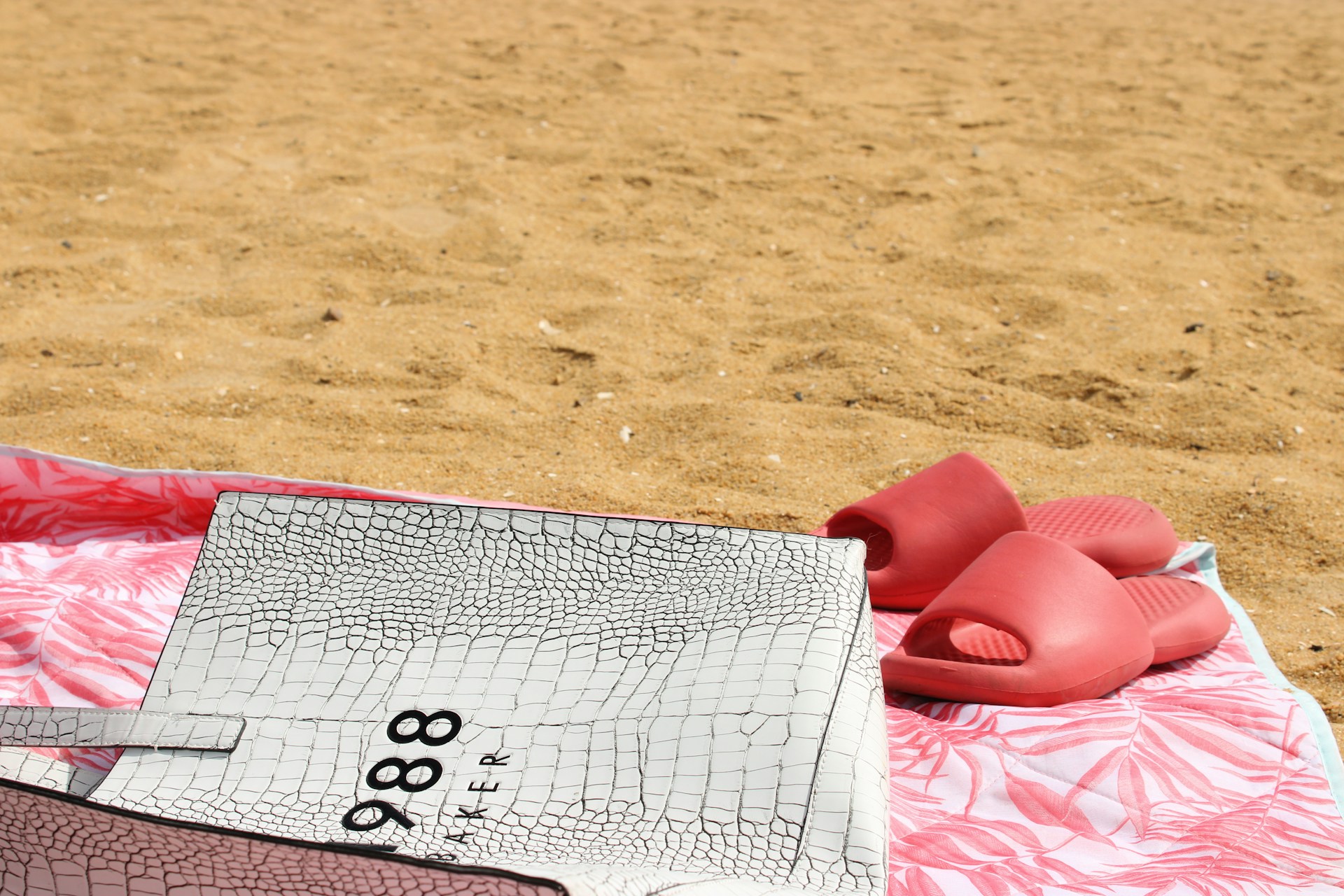 A sandy beach with a patterned white bag resting on a pink and white towel. A pair of red slides is placed next to the bag.