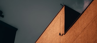 A geometric composition featuring part of a building with a warm, textured facade against a moody, dark sky. The contrast between the angular lines and the shadows creates a dramatic effect.