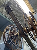 An intricate assembly of large, industrial gears and wheels is positioned beneath a corrugated metal roof. The machinery features metallic and brass components, with several cables and levers visible, suggesting a historical or industrial setting.