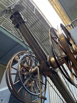 An intricate assembly of large, industrial gears and wheels is positioned beneath a corrugated metal roof. The machinery features metallic and brass components, with several cables and levers visible, suggesting a historical or industrial setting.