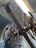 An intricate assembly of large, industrial gears and wheels is positioned beneath a corrugated metal roof. The machinery features metallic and brass components, with several cables and levers visible, suggesting a historical or industrial setting.