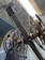 An intricate assembly of large, industrial gears and wheels is positioned beneath a corrugated metal roof. The machinery features metallic and brass components, with several cables and levers visible, suggesting a historical or industrial setting.