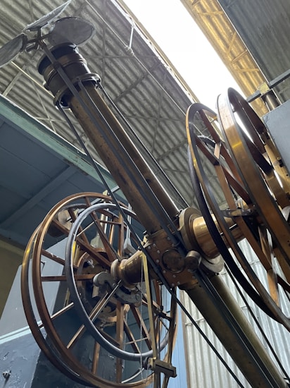 An intricate assembly of large, industrial gears and wheels is positioned beneath a corrugated metal roof. The machinery features metallic and brass components, with several cables and levers visible, suggesting a historical or industrial setting.