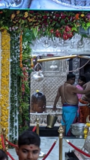 A sacred space adorned with a richly decorated floral garland hanging from the top. A large metallic idol or relic, possibly a religious artifact, is placed in the center. Several individuals, dressed in traditional attire, are performing some rituals with metal pots and other ceremonial items. The setting seems to be inside a temple or a religious shrine, with detailed carvings and embellishments visible in the background.