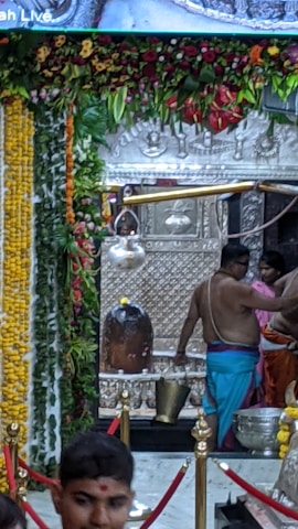 A sacred space adorned with a richly decorated floral garland hanging from the top. A large metallic idol or relic, possibly a religious artifact, is placed in the center. Several individuals, dressed in traditional attire, are performing some rituals with metal pots and other ceremonial items. The setting seems to be inside a temple or a religious shrine, with detailed carvings and embellishments visible in the background.