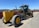 A large, yellow construction grader with the label 372GP is parked on a gravel surface. It has three axles with large tires and a long blade for grading soil. In the background, a red fence and a few scattered buildings are visible under a clear blue sky.