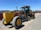 A large, yellow construction grader with the label 372GP is parked on a gravel surface. It has three axles with large tires and a long blade for grading soil. In the background, a red fence and a few scattered buildings are visible under a clear blue sky.