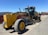 A large, yellow construction grader with the label 372GP is parked on a gravel surface. It has three axles with large tires and a long blade for grading soil. In the background, a red fence and a few scattered buildings are visible under a clear blue sky.
