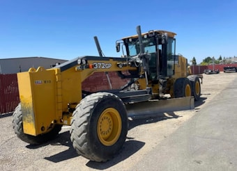 A large, yellow construction grader with the label 372GP is parked on a gravel surface. It has three axles with large tires and a long blade for grading soil. In the background, a red fence and a few scattered buildings are visible under a clear blue sky.
