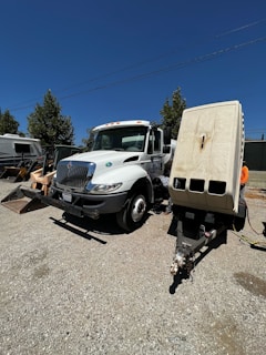 A large white utility truck is parked on a gravel surface next to a trailer with several vents or compartments. The scene is set outdoors under a clear blue sky, with trees and other vehicles in the background.