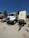 A large white utility truck is parked on a gravel surface next to a trailer with several vents or compartments. The scene is set outdoors under a clear blue sky, with trees and other vehicles in the background.
