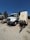 A large white utility truck is parked on a gravel surface next to a trailer with several vents or compartments. The scene is set outdoors under a clear blue sky, with trees and other vehicles in the background.