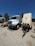 A large white utility truck is parked on a gravel surface next to a trailer with several vents or compartments. The scene is set outdoors under a clear blue sky, with trees and other vehicles in the background.