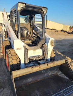 A compact skid-steer loader with large tires and a metal protective cage surrounding the driver's seat is parked on a dirt surface. The attachment at the front is a bucket, suggesting it is used for digging or transporting materials. The equipment is situated outdoors, with a clear blue sky in the background, and there are some storage containers or trailers nearby.